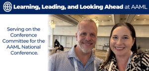 Two smiling people wearing conference lanyards take a selfie inside a large, well-lit event hall with chandeliers and round tables in the background.